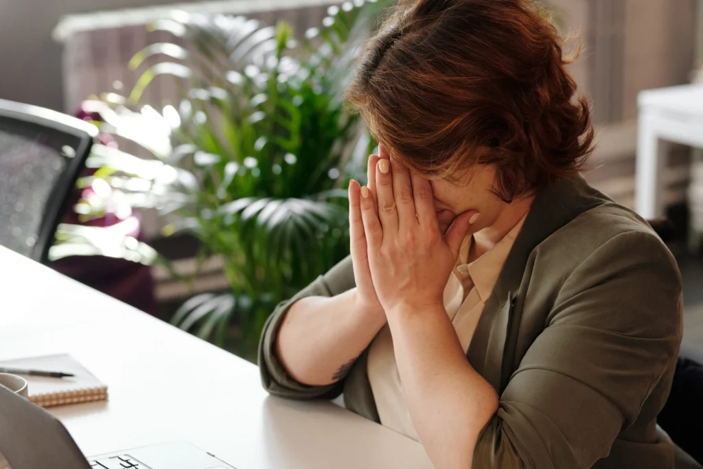 A professional woman in an office setting covering her face with her hands, depicting a moment of grief and emotional exhaustion at a desk with a laptop.